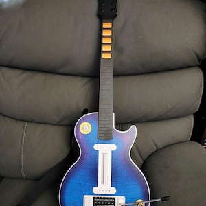 Puede incluir: Un controlador de guitarra Gibson azul y morado con mástil y diapasón negros. La guitarra tiene perillas doradas y un panel de control blanco. Una correa negra está sujeta a la guitarra.