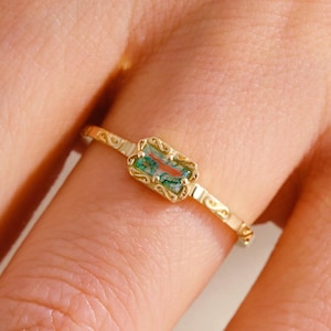 May include: A gold ring featuring a rectangular, green gemstone. The ring has a delicate, patterned band and a decorative setting around the gemstone. The ring is shown on a finger.