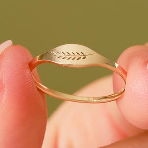 May include: A gold-colored ring with a leaf design. The ring is held between two fingers against a green background. The leaf design is etched into the ring's surface, adding a delicate detail to the jewelry.