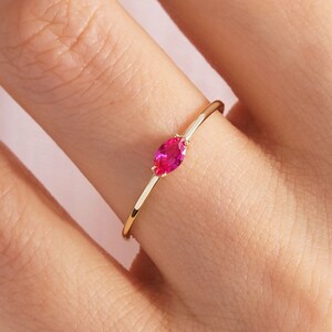 May include: A slender gold ring with a vivid oval-cut ruby gemstone. The ring's thin band enhances the bright red colour of the gemstone, creating a simple yet elegant design. The ring is shown on a person's finger.
