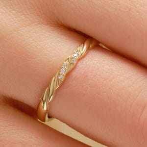 May include: A gold ring with a twisted rope design. The ring features small, clear gemstones. The ring is on a finger, with a blurred skin tone in the background.