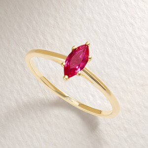 May include: A gold ring featuring a marquise-cut, vibrant red gemstone. The slender band is a polished gold, and the gemstone is set with delicate prongs. The ring is displayed on a neutral background, highlighting its elegant design.