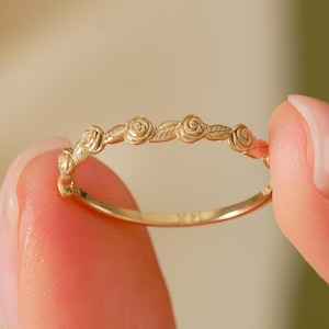May include: A delicate gold ring featuring a repeating floral design of small roses and leaves. The ring is held between two fingers, showcasing its intricate details and polished finish. The ring's band is thin, and the floral elements are evenly spaced.