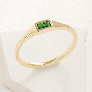 May include: A gold ring with a rectangular green gemstone set in a rectangular bezel. The ring is on a white surface and has a smooth, polished finish. The band is thin and rounded.
