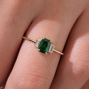 May include: A gold ring with a green emerald gemstone and two smaller baguette-cut diamonds.