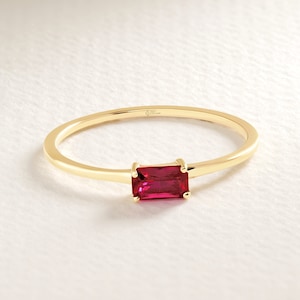 May include: A gold ring featuring a rectangular, deep red gemstone. The slender band holds a horizontally set gemstone at the centre. The ring is presented on a white surface, accentuating its elegant design. The gemstone is approximately 5mm x 3mm.