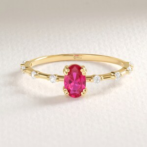 May include: A delicate gold ring featuring an oval-cut, vibrant pink gemstone as the centerpiece. The band is adorned with small, sparkling white gemstones and a textured design. The ring is set against a soft, neutral background, highlighting its elegant design.