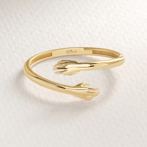 May include: A gold ring with two hands clasped together, forming a circle. The ring is open and can be adjusted to fit different finger sizes.