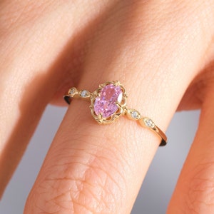 May include: A delicate gold ring with a pink oval gemstone set in a filigree design. The ring is adorned with small diamonds on the band.