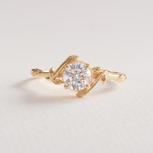 May include: A gold engagement ring with a large, round, clear gemstone. The ring features a nature-inspired design with delicate leaf and vine details. The band and setting are a warm gold color, creating an elegant and timeless look.