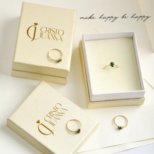 K&ouml;nnte beinhalten: Drei wei&szlig;e Schachteln mit goldenen Schriftz&uuml;gen, die "Cristo Juana" und den Spruch "make happy be happy" lesen. Jede Schachtel enth&auml;lt einen goldenen Ring mit einem gr&uuml;nen Edelstein.