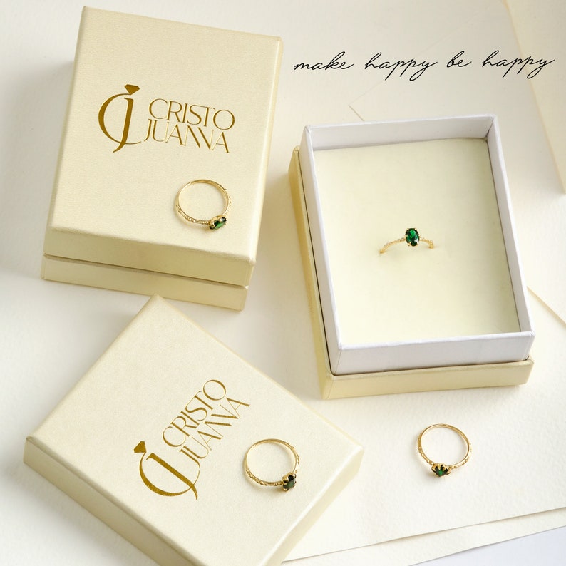 K&ouml;nnte beinhalten: Drei wei&szlig;e Schachteln mit goldenen Schriftz&uuml;gen, die "Cristo Juana" und den Spruch "make happy be happy" lesen. Jede Schachtel enth&auml;lt einen goldenen Ring mit einem gr&uuml;nen Edelstein.