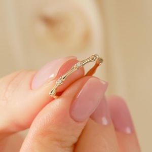 May include: A thin gold ring featuring a bamboo-inspired design with segmented nodes. The band is accented with three small, clear, round-cut stones set at intervals along the top surface, creating a delicate and polished metallic appearance.