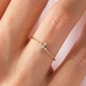 May include: A delicate gold ring featuring a small diamond cross design. The ring is worn on a finger.