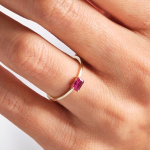 May include: A gold ring with a rectangular, bright pink gemstone. The ring's thin band curves elegantly, accentuating the vivid gemstone. The ring is displayed on a finger, highlighting its refined design.