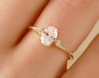 May include: A gold engagement ring featuring an oval-cut diamond as the centerpiece. The ring has a delicate band with small accent diamonds on each side, creating a vintage-inspired design. The ring is shown on a person's finger.
