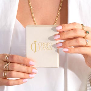 以下が含まれることがあります： 「CRISTO JUANA」の金色の文字が入ったクリーム色の箱を持つ人。その人は複数の金の指輪と金のチェーンを身につけています。その人はピンクのマニキュアをしています。
