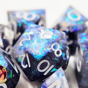 May include: A set of iridescent dice with black and white numbers. The dice are made of resin and have a glittery finish. The dice are in a variety of shapes, including a d20, d12, d10, d8, d6, and d4.