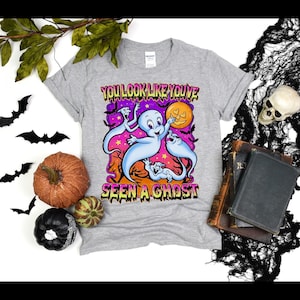 Könnte beinhalten: Ein graues T-Shirt mit einem Cartoon-Geister-Design und dem Text "YOU LOOK LIKE YOU'VE SEEN A GHOST". Das Design zeigt einen lächelnden Geist, einen Vollmond und andere Geister. Das T-Shirt ist von Halloween-Dekorationen umgeben.