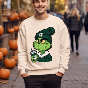May include: Cream-colored sweatshirt featuring a green Grinch character wearing a green beanie with a white and green logo. The Grinch is holding a coffee cup with a green and white logo. The sweatshirt has a crew neck and long sleeves.
