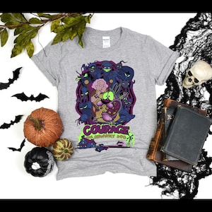 Puede incluir: Una camiseta gris claro con un gráfico de la serie animada, Coraje, el perro cobarde. El diseño incluye a Coraje, Muriel y varios personajes espeluznantes en tonos morados, verdes y azules. La camiseta tiene el logo del programa.