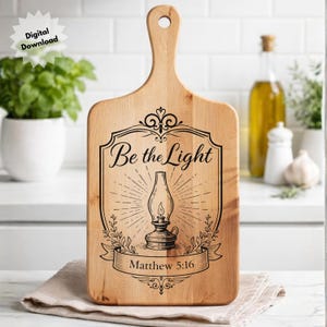 May include: A light-colored wooden cutting board with a handle. The board features a black graphic of a lamp with the text "Be the Light" and "Matthew 5:16". The board is rectangular with a rounded top and a hole for hanging.
