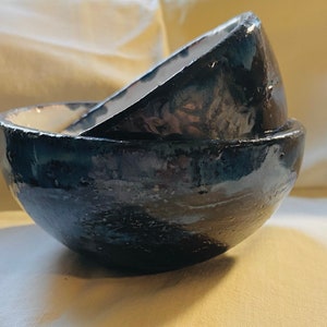 May include: Two ceramic bowls, one nested inside the other. The bowls are a dark blue color with a glossy finish. The bowls have a slightly irregular shape and a white rim.