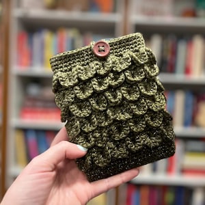 May include: A handmade, olive green crochet sleeve with a scale-like pattern. The sleeve is secured with a small, round, brown button. The background is a blurred bookshelf, suggesting a book cover or e-reader case.