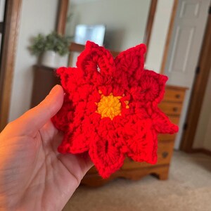 May include: A handmade, red crochet poinsettia flower with a yellow center. The flower has multiple petals and is held in a hand. The background includes a wooden dresser and a mirror.