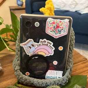 May include: A black smartphone with stickers, including one that says "Ask me for book recs" and another with a floral design. The phone is in a green crocheted case, with a yellow crocheted accent at the top. The words "Currently Reading" are also visible.