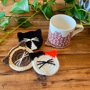 May include: A white mug with a red pattern sits on a small, round, woven coaster. Three handmade crochet cat face coasters in black, brown, and white are arranged on a wooden surface. Green leaves and a plant are in the background.