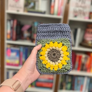 Puede incluir: Una funda de teléfono de ganchillo hecha a mano en tonos de azul, verde, amarillo y marrón. La funda presenta un gran diseño de girasol en la parte delantera. La funda se sostiene frente a una estantería.