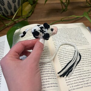 Könnte beinhalten: Handgefertigter Häkel-Kuh-Lesezeichen mit schwarzen Flecken, einer rosa Nase und einem weißen Körper. Das Lesezeichen befindet sich auf einem Buch mit Text. Der Schwanz des Lesezeichens ist schwarz und weiß.