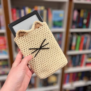 May include: A beige crochet Kindle case shaped like a cat, with brown ears and black whiskers. The case is holding an e-reader. The background is a bookshelf filled with books.