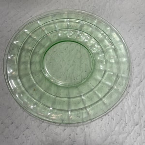 May include: A round, light green glass plate featuring a smooth, circular centre surrounded by a wide, textured rim with vertical, radiating ridges. The translucent glass has a subtle, uniform pale green tint throughout its entire surface.
