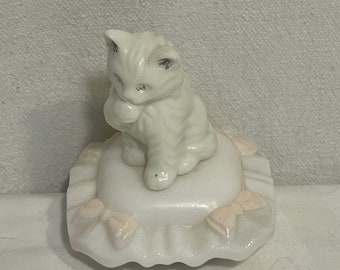 Puede incluir: Una figura de cerámica blanca de un gatito sentado sobre un cojín cuadrado con volantes. El gatito tiene detalles grises sutiles en las orejas y la cara. El cojín presenta lazos de color rosa pálido en cada esquina y un borde con volantes.