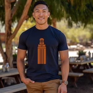 May include: Navy blue t-shirt with a burnt orange graphic of a tower. The short-sleeved shirt is made of a soft material and is being worn outdoors. The person is wearing a watch and khaki pants.