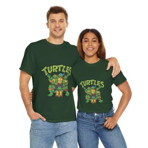 Könnte beinhalten: Zwei Personen tragen passende dunkelgrüne T-Shirts mit einer Grafik der Teenage Mutant Ninja Turtles. Die Shirts haben das Wort "TURTLES" in einer gelben, stilisierten Schriftart über den Zeichentrickfiguren.