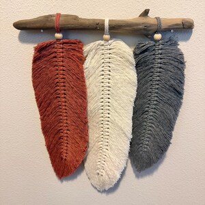 May include: Three macrame feathers in shades of rust, cream, and gray hang from a piece of driftwood. Each feather is suspended by a cord and wooden bead. The feathers are arranged in a row, creating a rustic wall hanging.