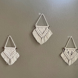 May include: Three white macrame wall hangings with wooden dowels, each with a different pattern, hanging on a gray wall.