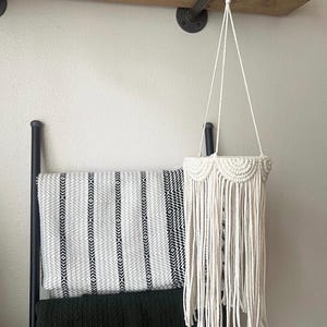 May include: A cream-colored macrame plant hanger featuring a series of semi-circular knotted patterns along the top rim and long, loose fringe hanging downward. The hanger is suspended from a wooden ring attached to a black metal pipe fixture.