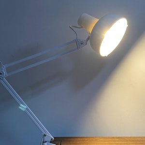 May include: A white adjustable desk lamp with a bright light, mounted on a wooden desk. The lamp's arm is articulated, allowing for flexible positioning. The lamp is against a blue-gray wall.