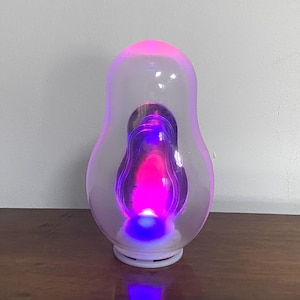 Könnte beinhalten: Dekorative Lampe mit einer klaren, bauchigen Außenschale. Im Inneren leuchtet eine kleinere, birnenförmige Form mit leuchtendem rosa, lila und blauem Licht. Die Lampe steht auf einem weißen Sockel und strahlt eine sanfte, farbenfrohe Beleuchtung aus.