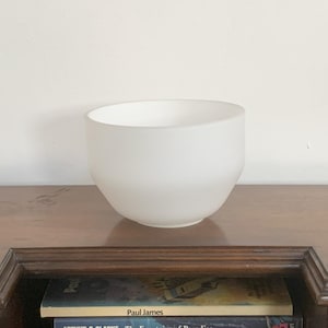 May include: A white frosted glass bowl on a wooden surface. The bowl is round and has a smooth, matte finish.