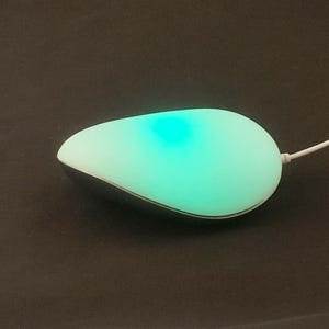以下が含まれることがあります： 下からターコイズブルーの光を放つ白いワイヤレスマウス。マウスは涙の形をしており、コードが接続されています。