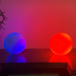 Puede incluir: Dos orbes brillantes, uno azul y otro rojo, se encuentran sobre una superficie oscura. Los orbes están iluminados desde adentro, proyectando un suave resplandor.