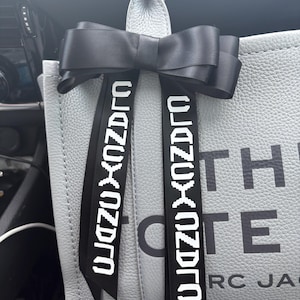 May include: A gray tote bag with a black bow and black straps. The straps have the text "0123456789" printed on them. The bag has the text "THE" and "OTE" printed on it. The text "MARC JAC" is partially visible at the bottom of the bag.