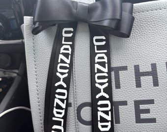 May include: A gray tote bag with a black bow and black straps. The straps have the text "0123456789" printed on them. The bag has the text "THE" and "OTE" printed on it. The text "MARC JAC" is partially visible at the bottom of the bag.