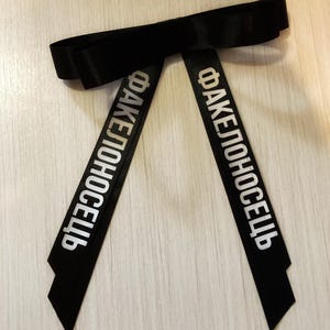 May include: Black ribbon bow with the Russian text "FAKEЛОНОСЕЦЬ" printed in white. The ribbon is folded into a bow shape at the top, with two long, angled tails extending downwards. The ribbon is lying on a light wood-grain surface.