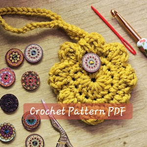 Può includere: Primo piano di un fiore all'uncinetto giallo con un bottone al centro e un anello. Diversi bottoni decorativi con motivi colorati sono sparsi intorno al fiore. Sono visibili anche un uncinetto, un ago e delle forbici. Il testo "Crochet Pattern PDF" è sovrapposto all'immagine.
